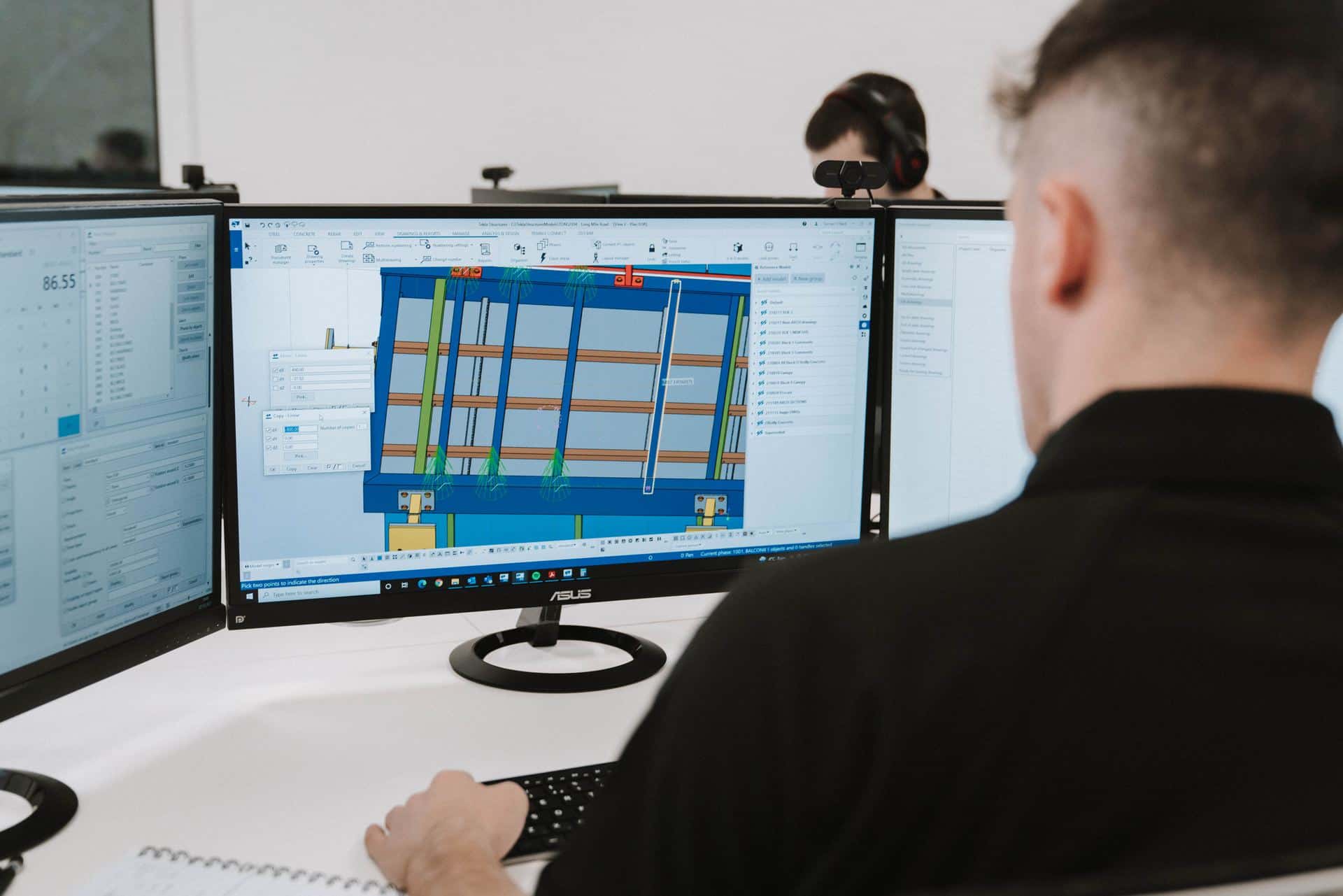 Two OD3 engineers at multi-monitor Tekla workstations, the Long Mile Road balcony model open on the near screen
